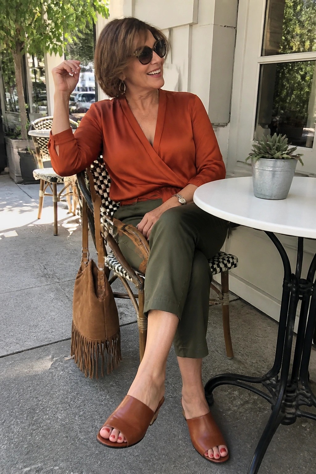 Woman over 50 wearing a rust orange wrap blouse tucked into olive green cropped pants with brown leather slide sandals and a brown suede fringed bag seated at an outdoor table.