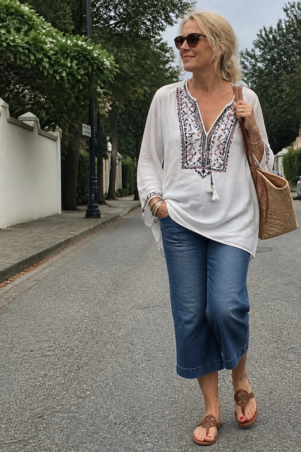 Woman over 50 in white embroidered boho tunic top with blue wide cropped jeans, brown thong sandals, woven straw tote bag, sunglasses, and stacked bracelets standing on a paved street.
