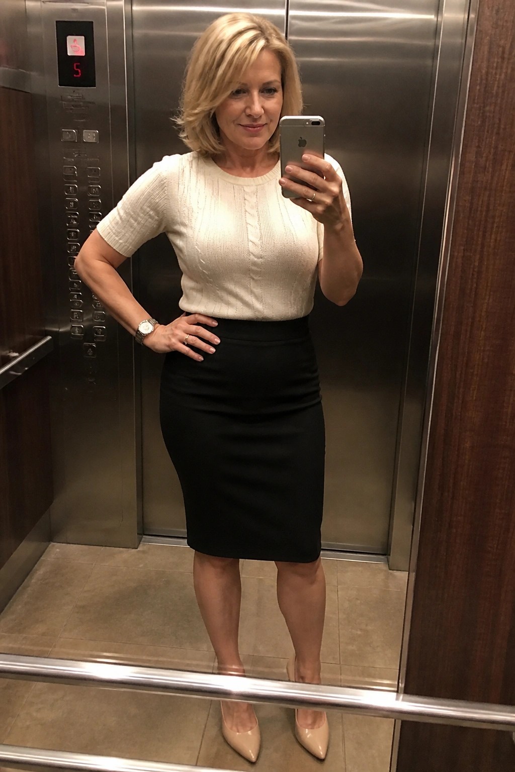 Blonde woman in white top and black skirt taking mirror selfie in elevator
