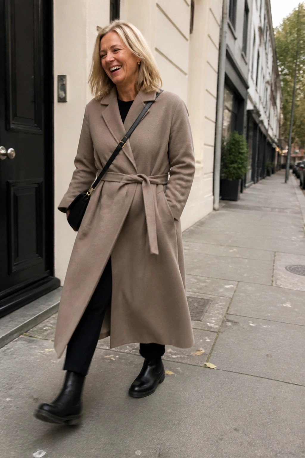 Smiling blonde woman in belted beige coat and black boots on city street.