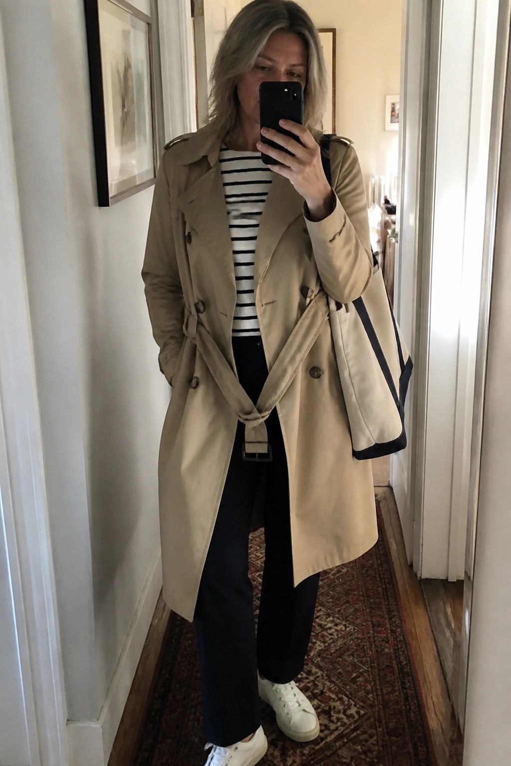 Woman over 50 in a beige trench coat belted at the waist over a black and white striped shirt paired with dark pants and white sneakers while holding a black phone for a mirror selfie and carrying a matching tote bag.