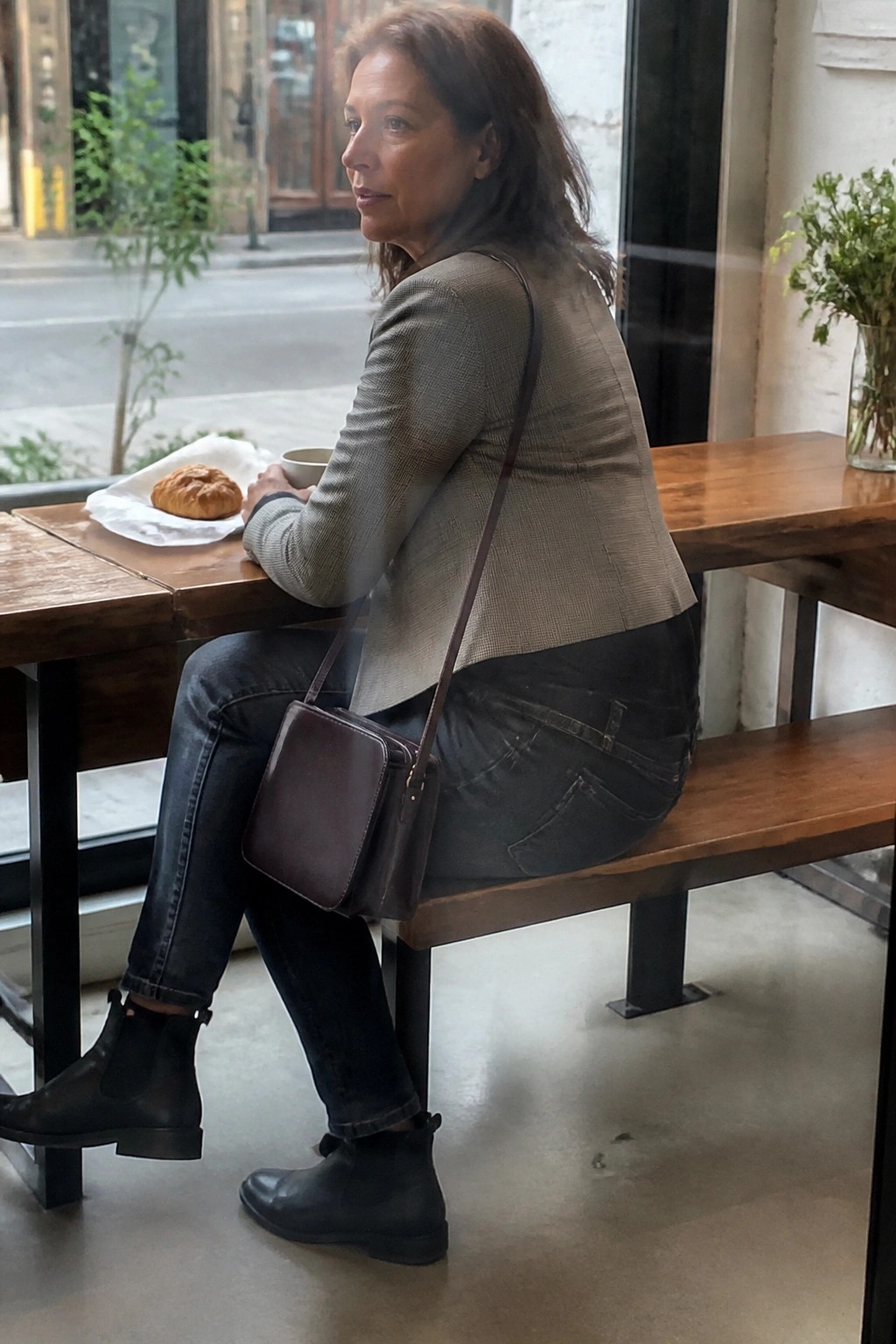 Woman in gray jacket and jeans at wooden cafe table with croissant and coffee