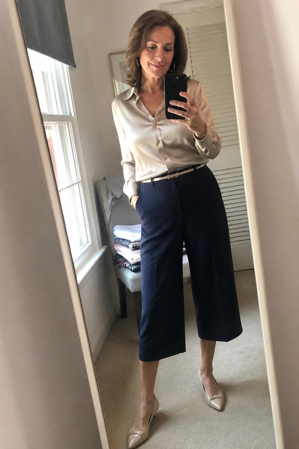 Woman in satin blouse and navy culottes taking mirror selfie with smartphone