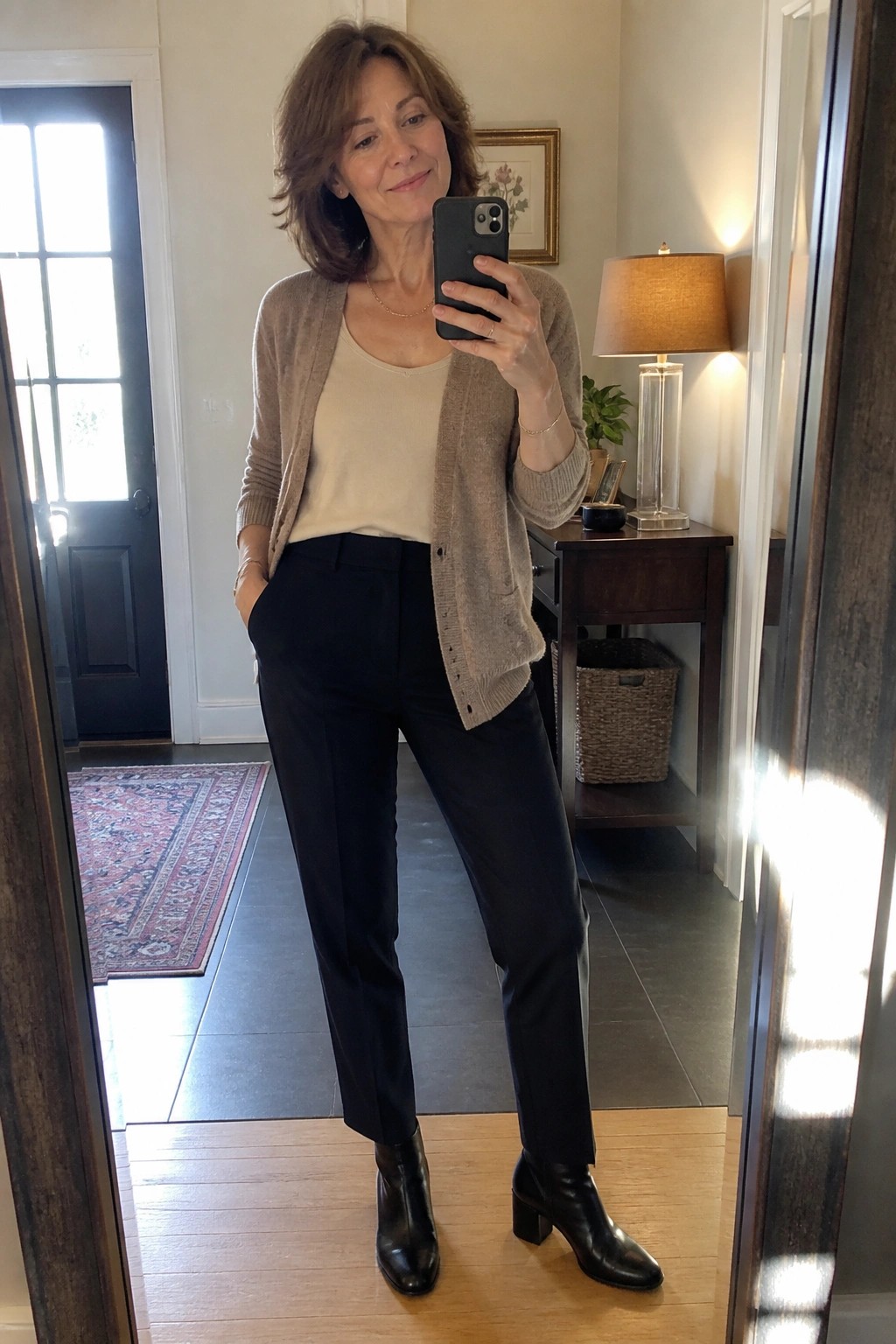 Woman over 50 wearing a taupe knit cardigan over a cream tank top with black tailored pants and black ankle boots in a mirror selfie.