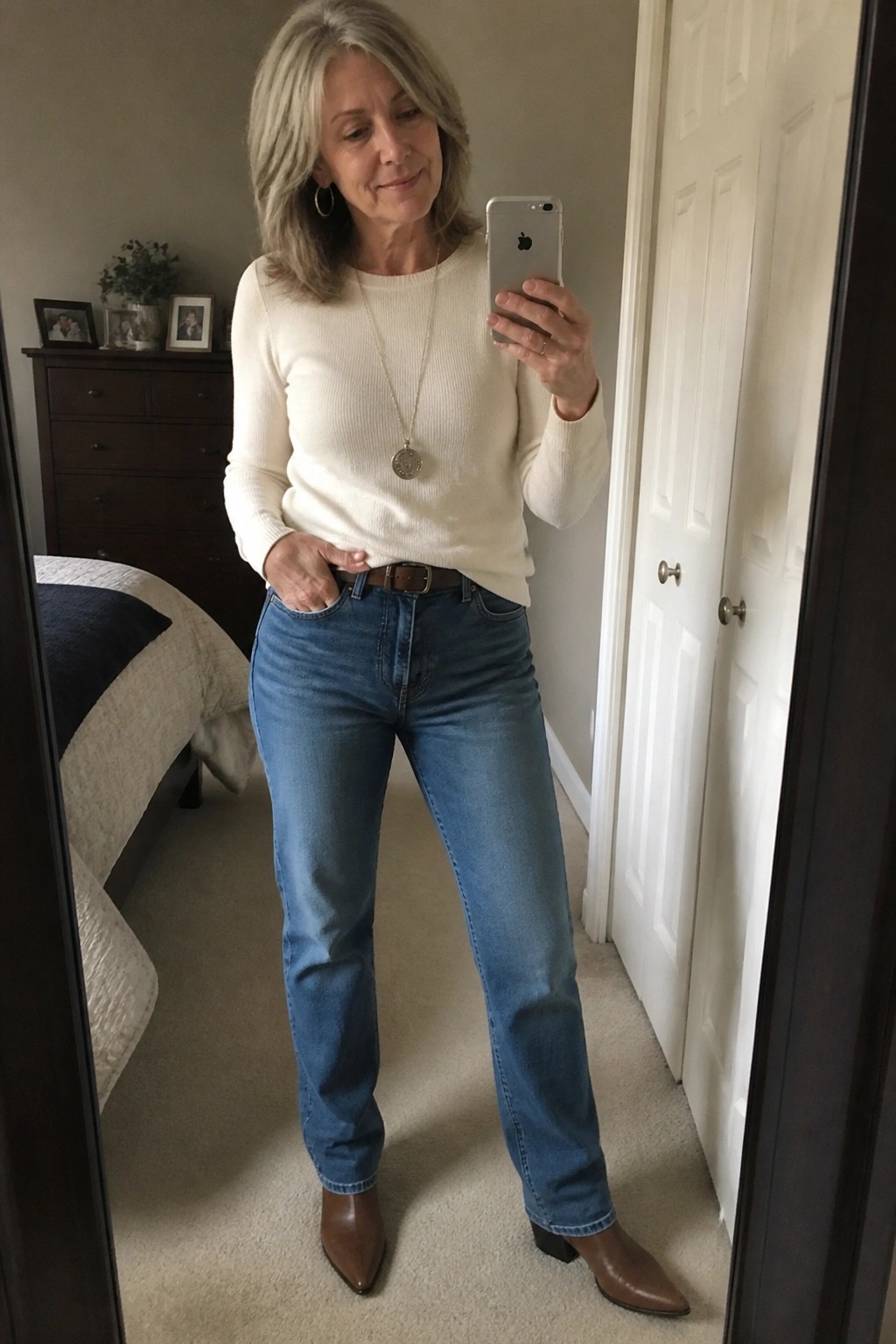 Woman with gray hair in cream sweater and blue jeans taking bedroom mirror selfie