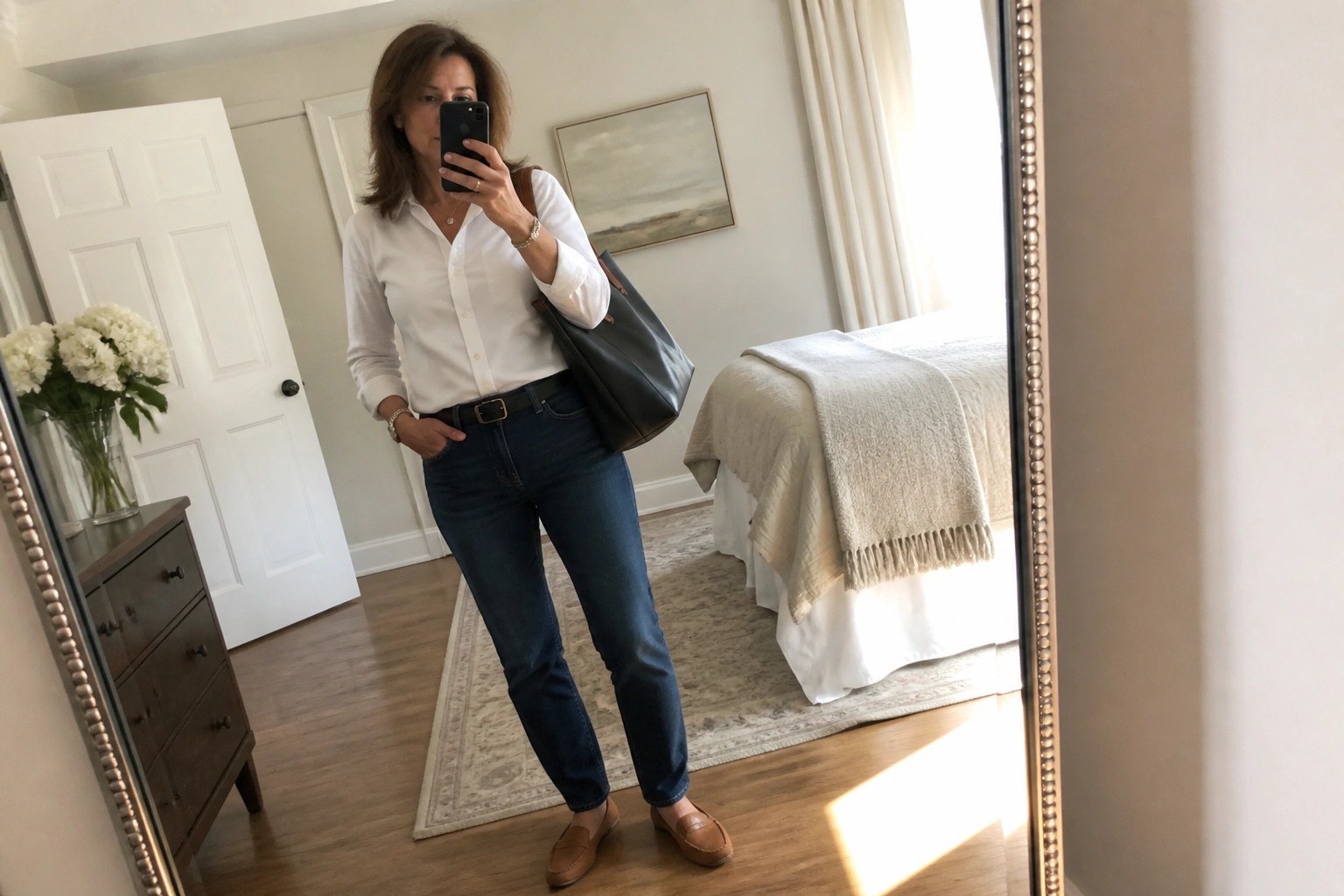 Woman in white shirt and blue jeans taking mirror selfie in bedroom with handbag