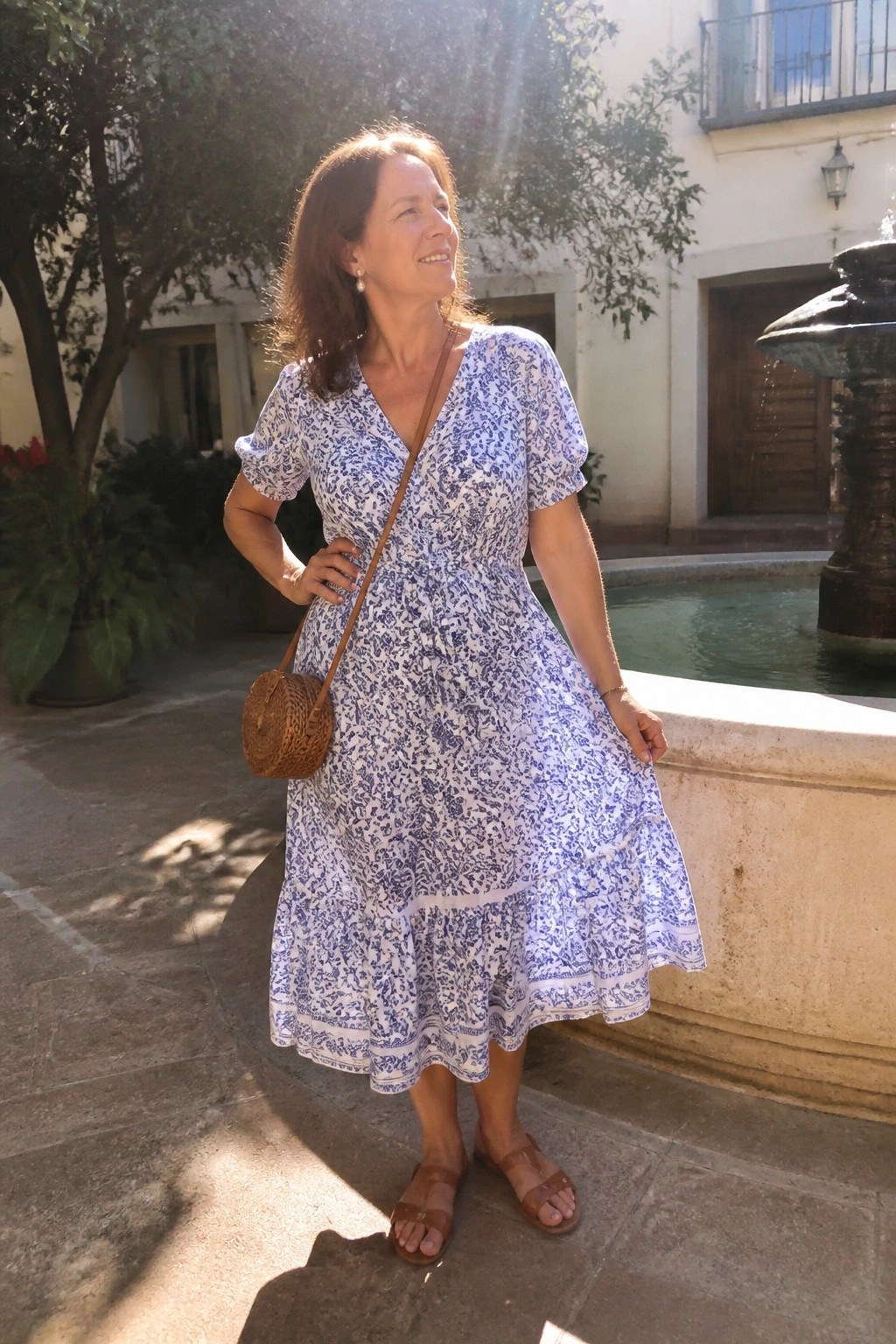 Woman in blue floral dress with woven bag standing by stone fountain