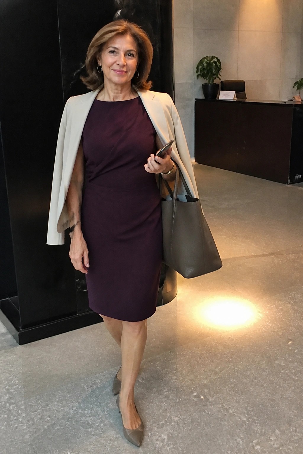 Woman in maroon dress with beige blazer holds phone and gray bag in lobby