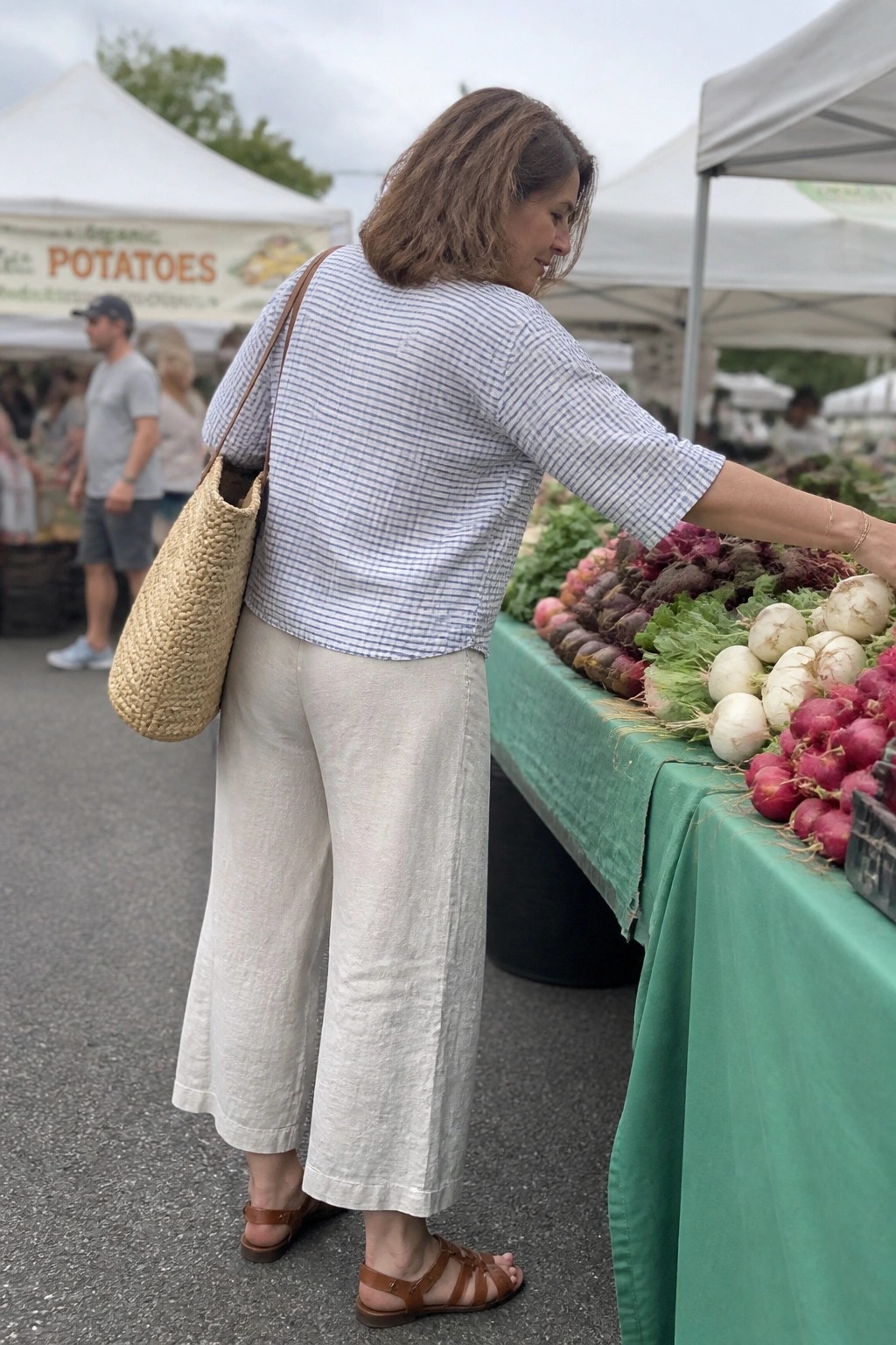 Woman in striped top and linen pants reaches for vegetables at market stall