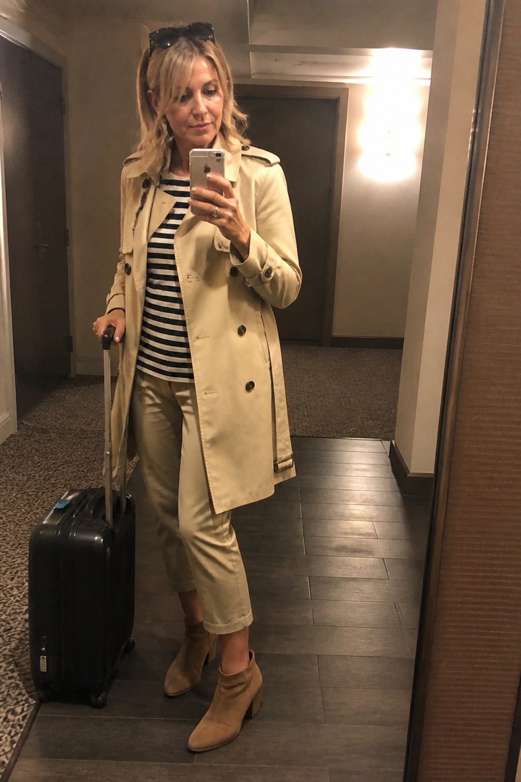 Blonde woman in beige trench coat holding black suitcase takes mirror selfie in hallway
