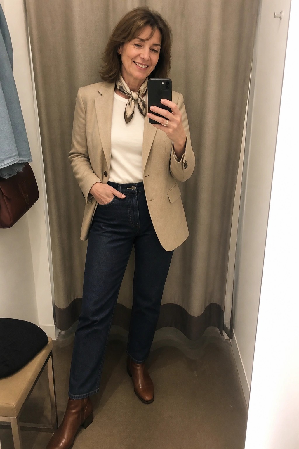 Woman in beige blazer, scarf, white top, jeans, and brown boots taking mirror selfie