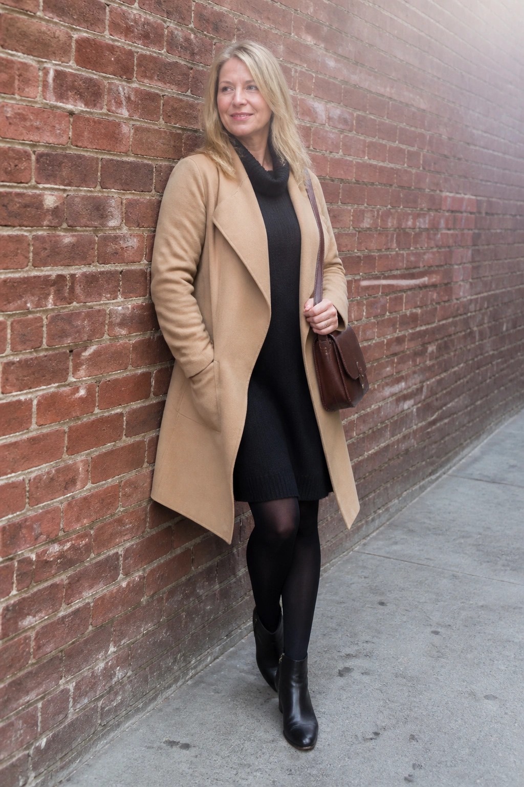 Woman in tan coat over black dress leaning on brick wall holding brown bag