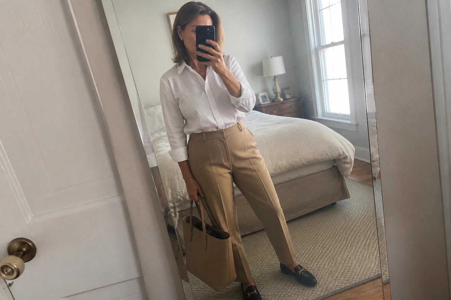 Woman in white shirt and tan pants holding brown handbag in bedroom mirror selfie