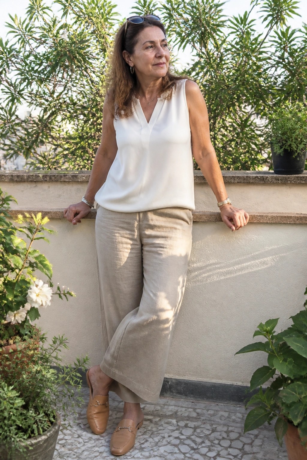 Woman in white sleeveless top and beige wide pants leaning on ledge amid potted plants.