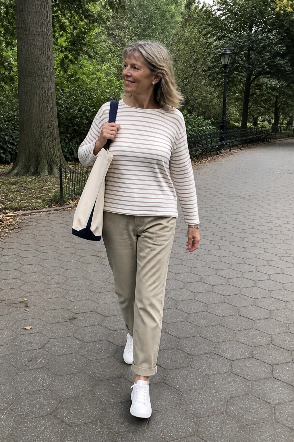 Smiling woman in striped top and beige pants carrying tote bag on park path