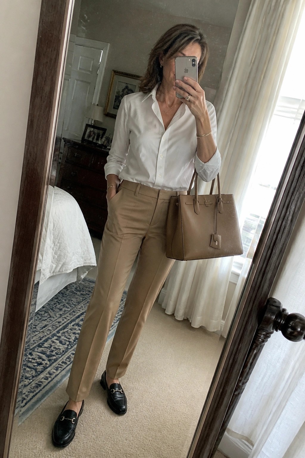 Woman in white shirt and tan pants holding brown handbag in bedroom mirror selfie