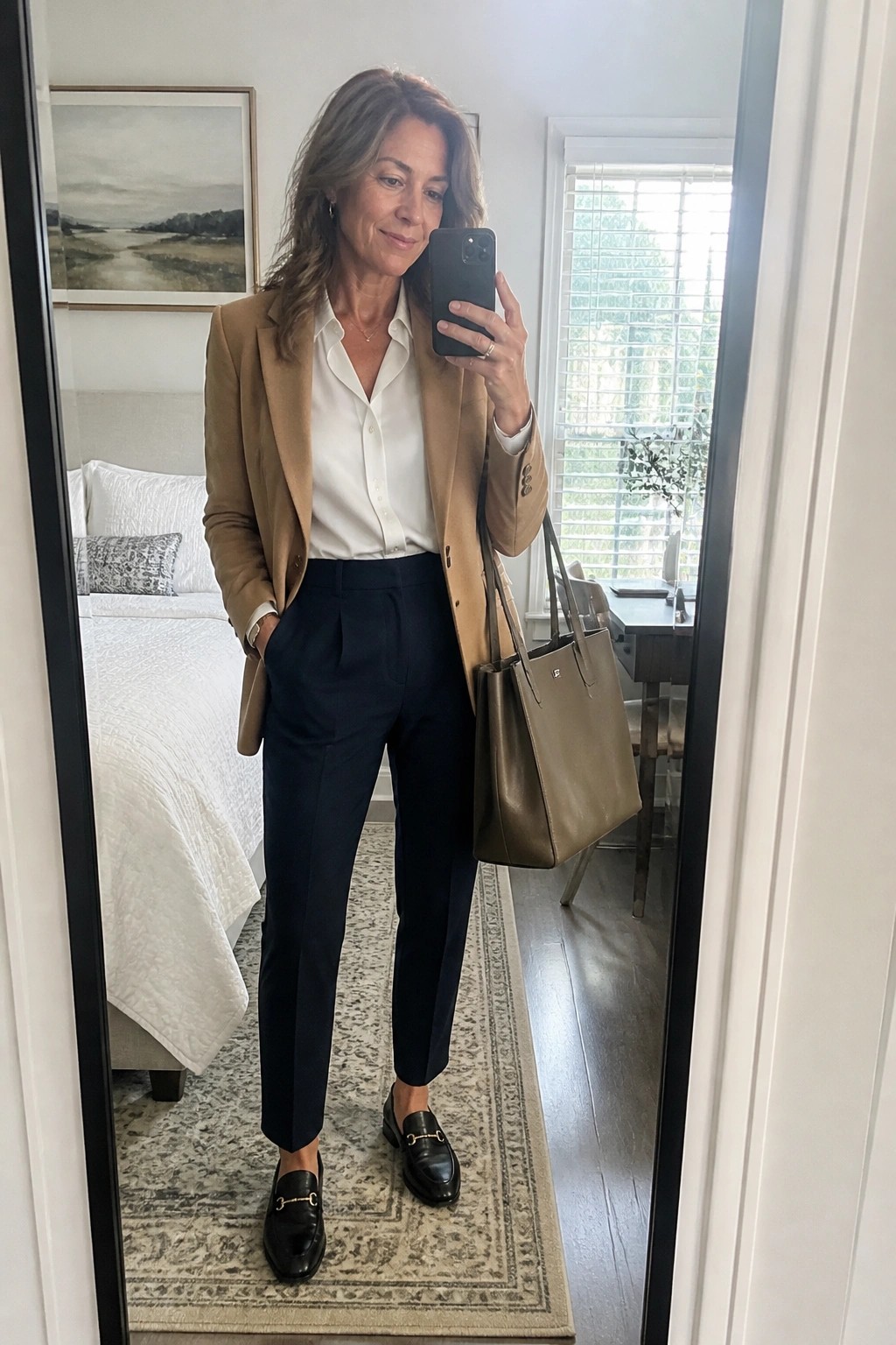 Woman in tan blazer and navy pants taking mirror selfie with leather tote bag