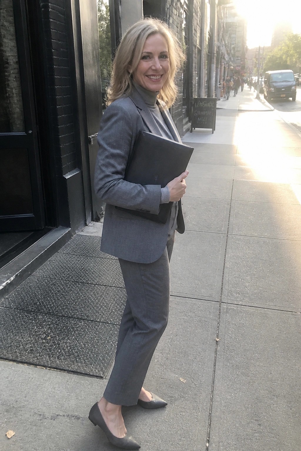 Smiling blonde woman in gray suit holding black folder on city sidewalk