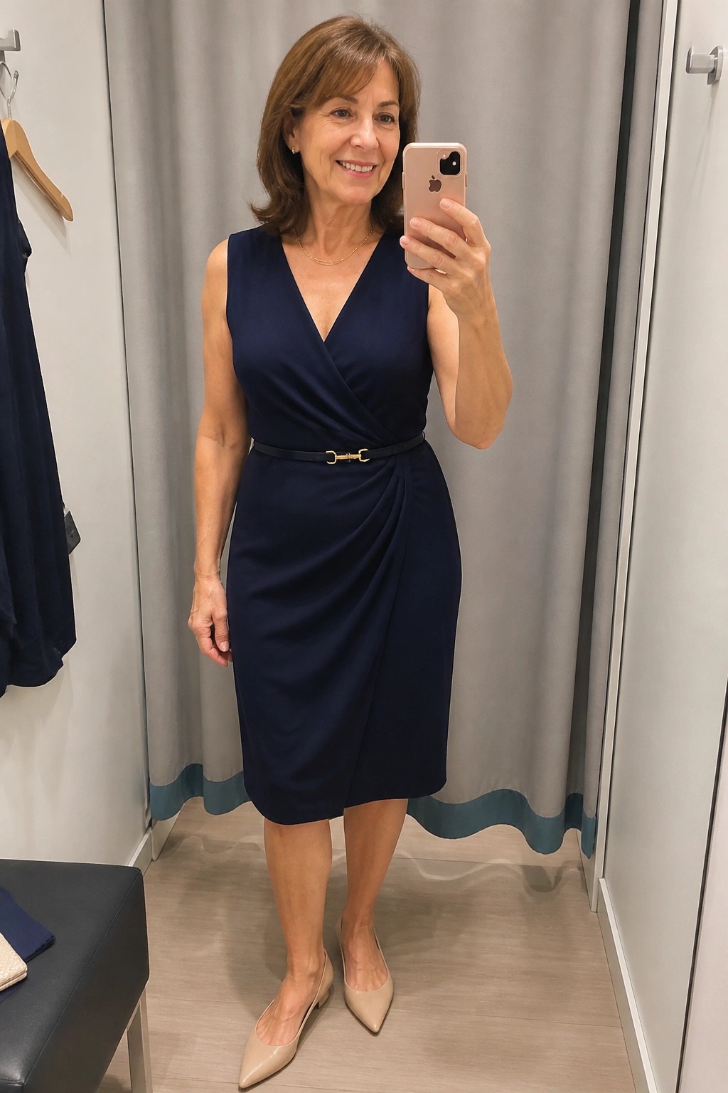 Woman in navy wrap dress taking mirror selfie with pink phone in fitting room