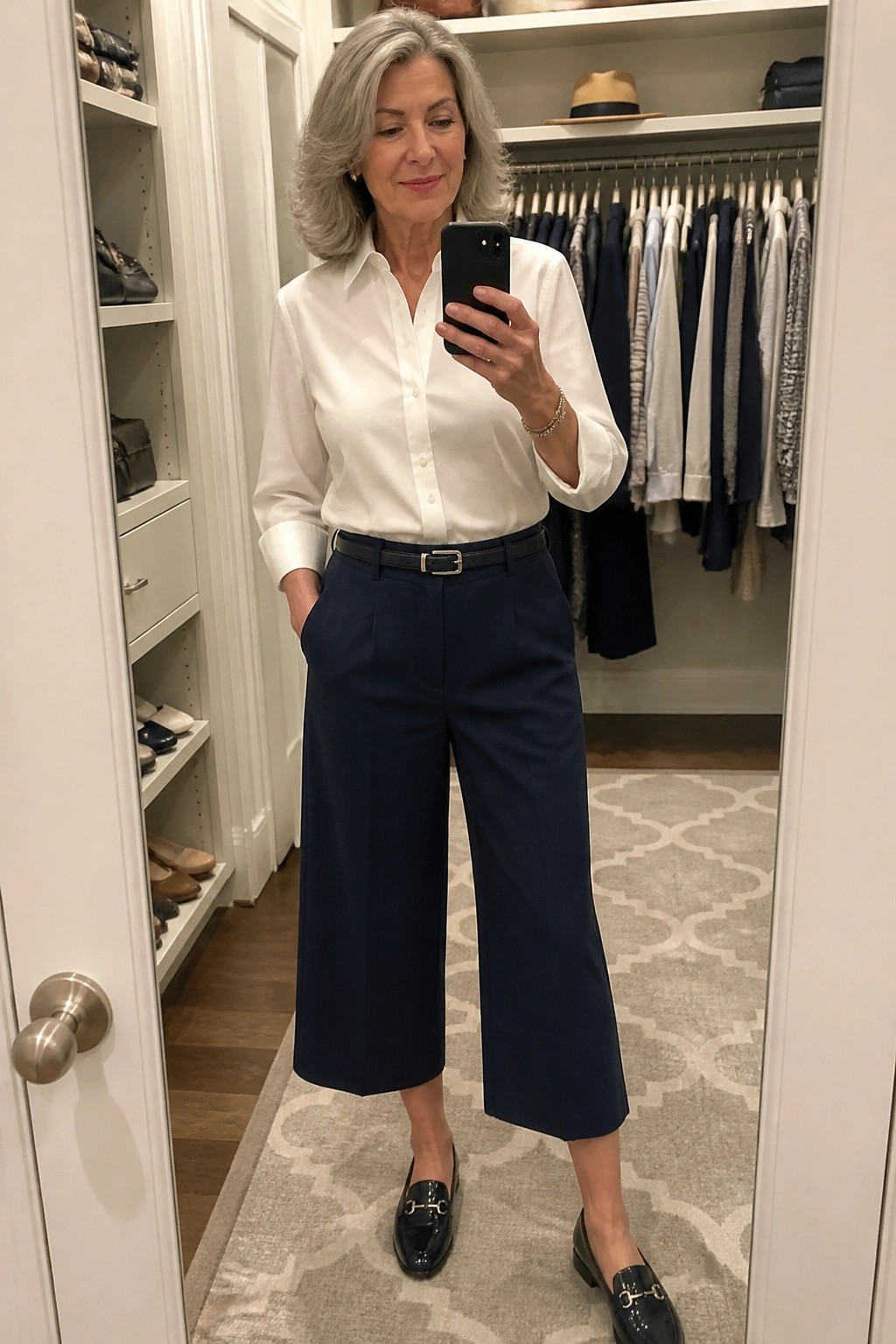 Gray-haired woman in white shirt and navy cropped pants taking closet mirror selfie