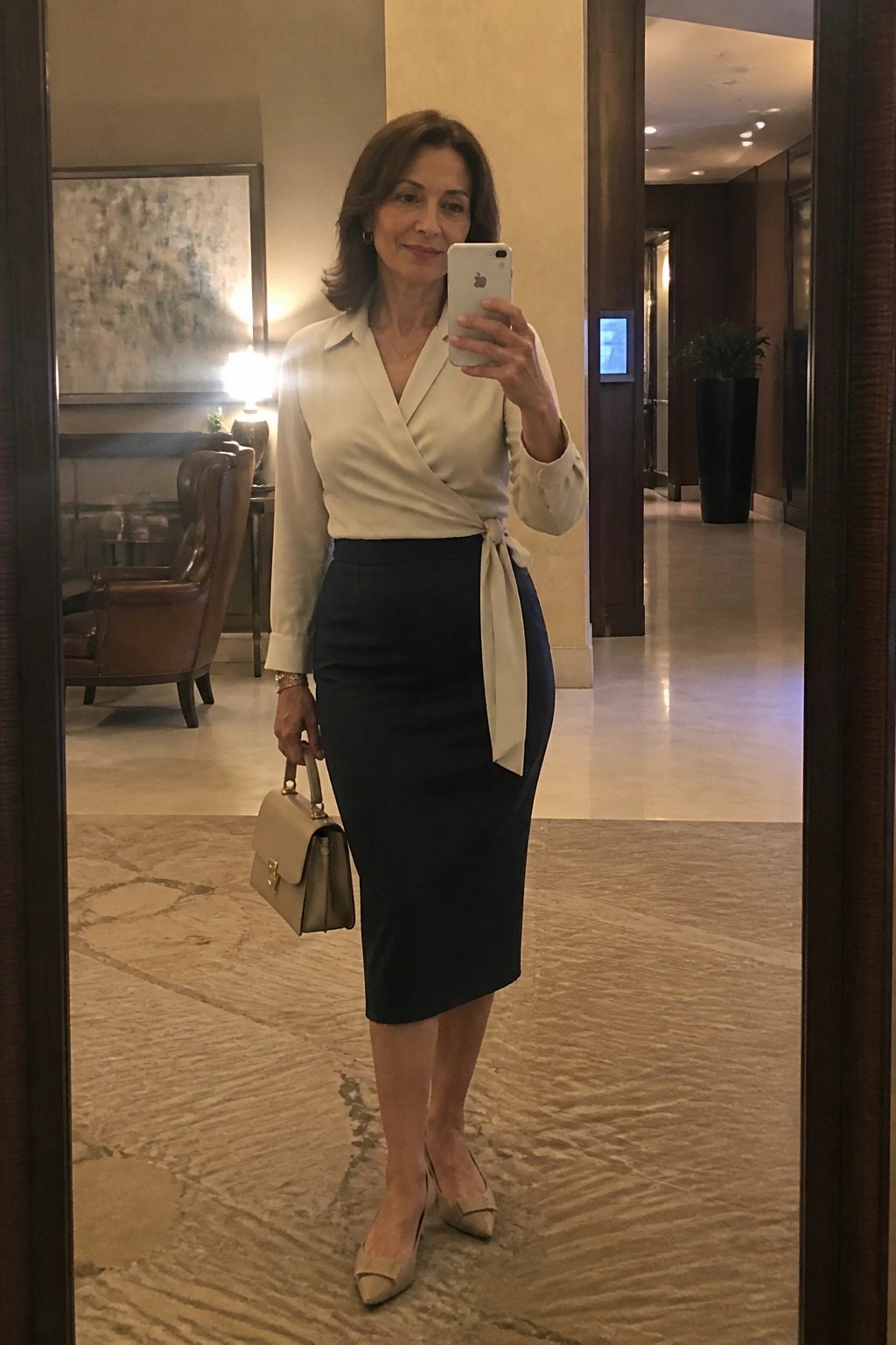 Woman in white wrap blouse and navy skirt holding phone and handbag in lobby.