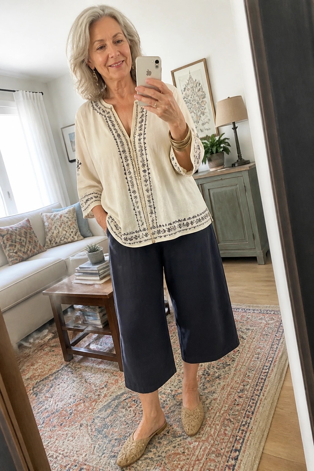 Woman with gray hair in cream embroidered top and navy culottes taking mirror selfie