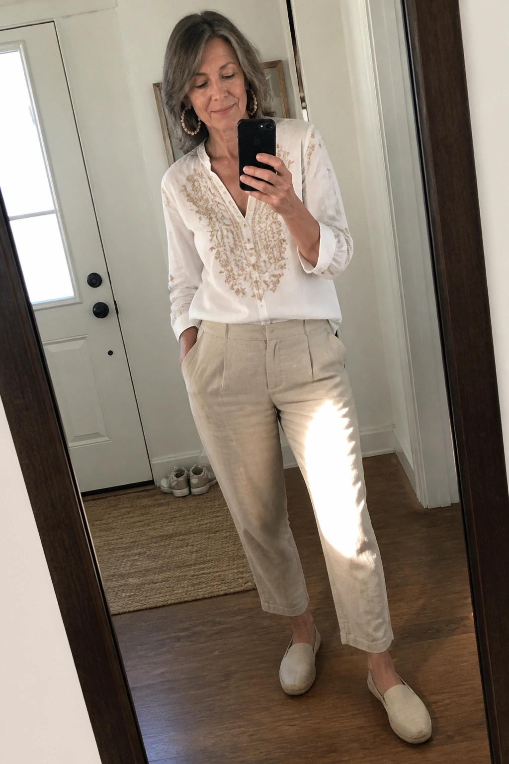 Gray-haired woman in white embroidered blouse and beige pants taking mirror selfie in hallway.