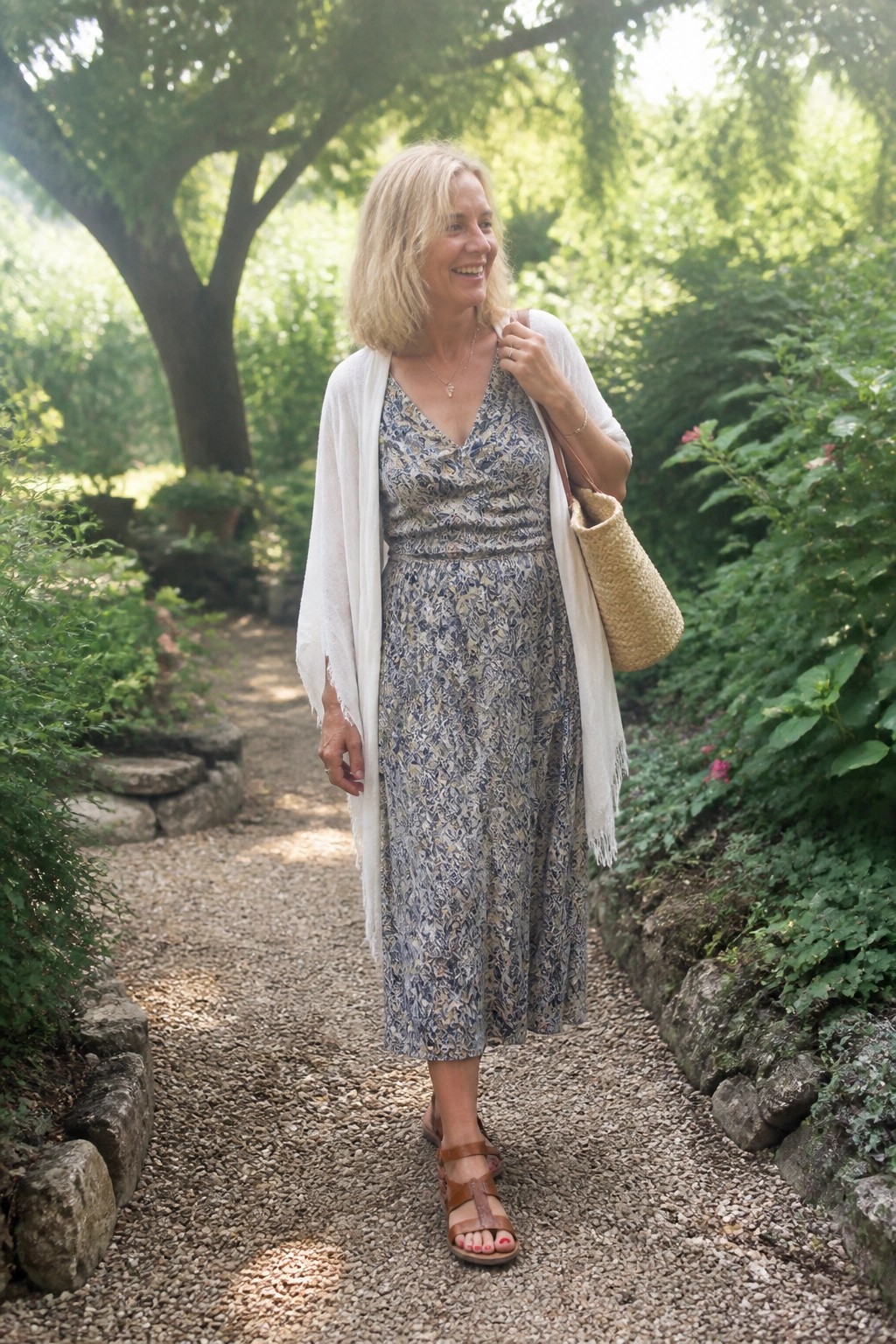 Blonde woman in floral dress and white shawl on sunny garden path with straw bag