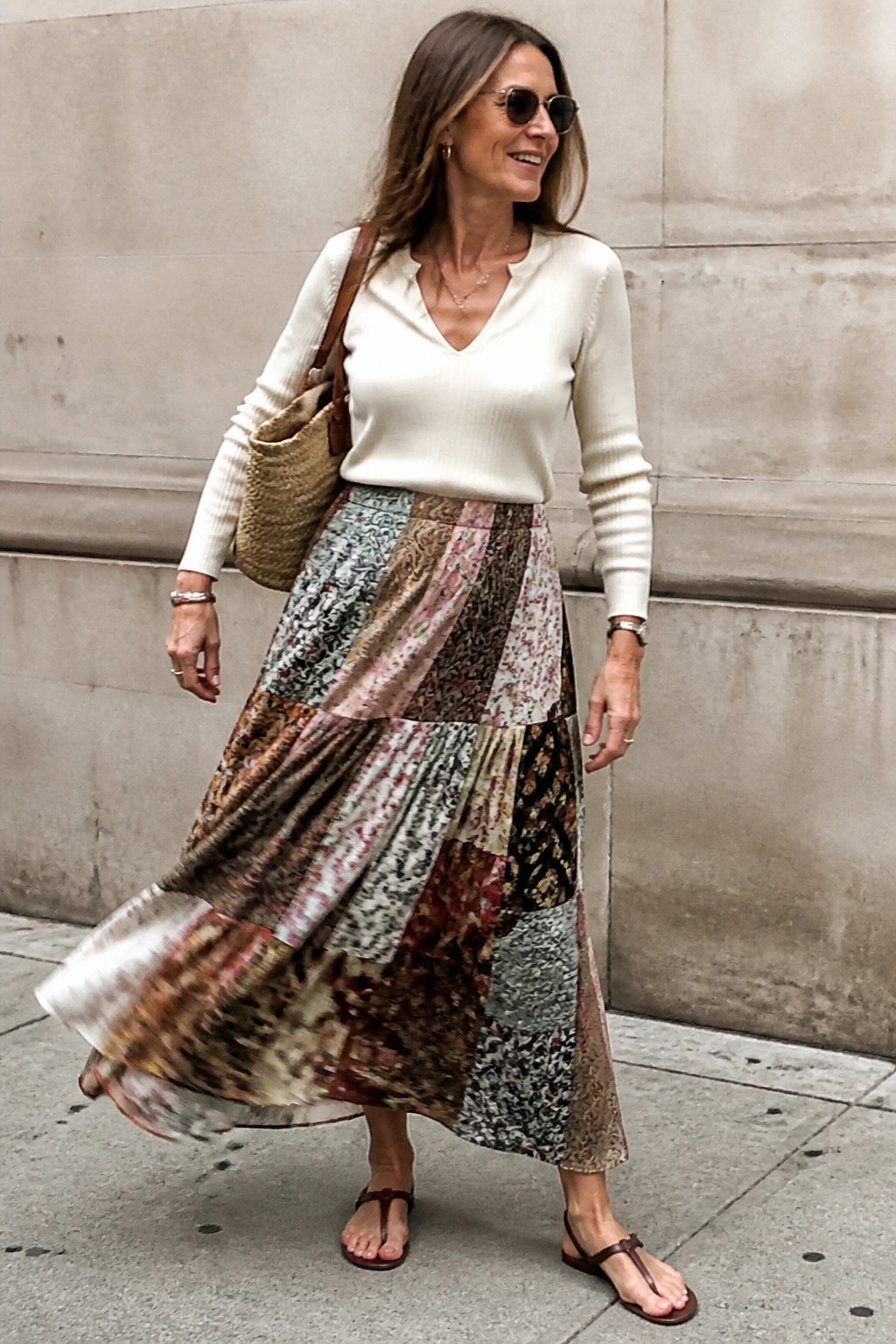 Woman over 50 in cream ribbed v-neck sweater tucked into a tiered patchwork maxi skirt with mixed floral prints, paired with brown thong sandals and a woven straw bag over one shoulder.