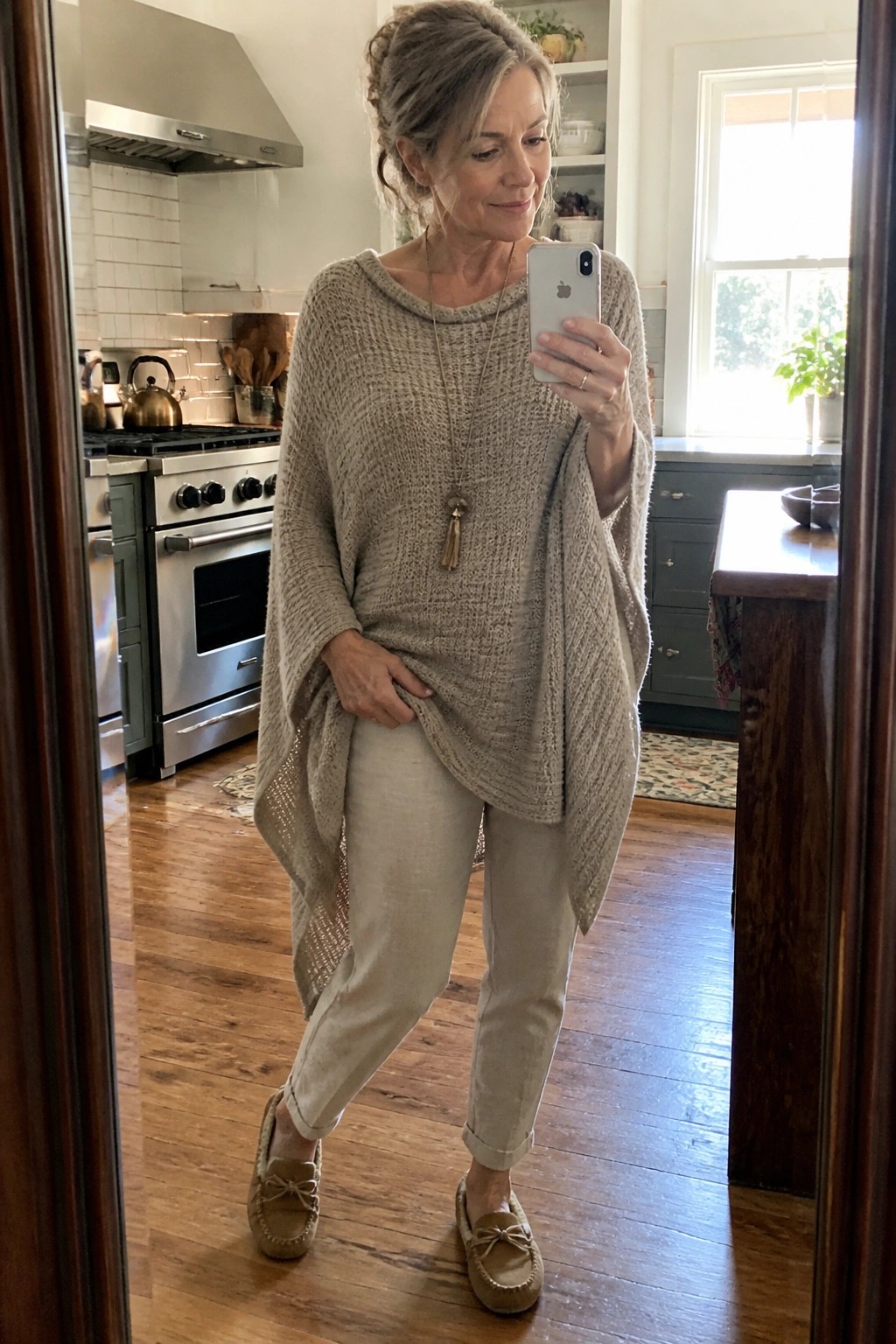 A woman over 50 wearing a beige knitted poncho sweater over light beige pants and matching moccasins, accessorized with a long necklace, taking a mirror selfie in a kitchen.