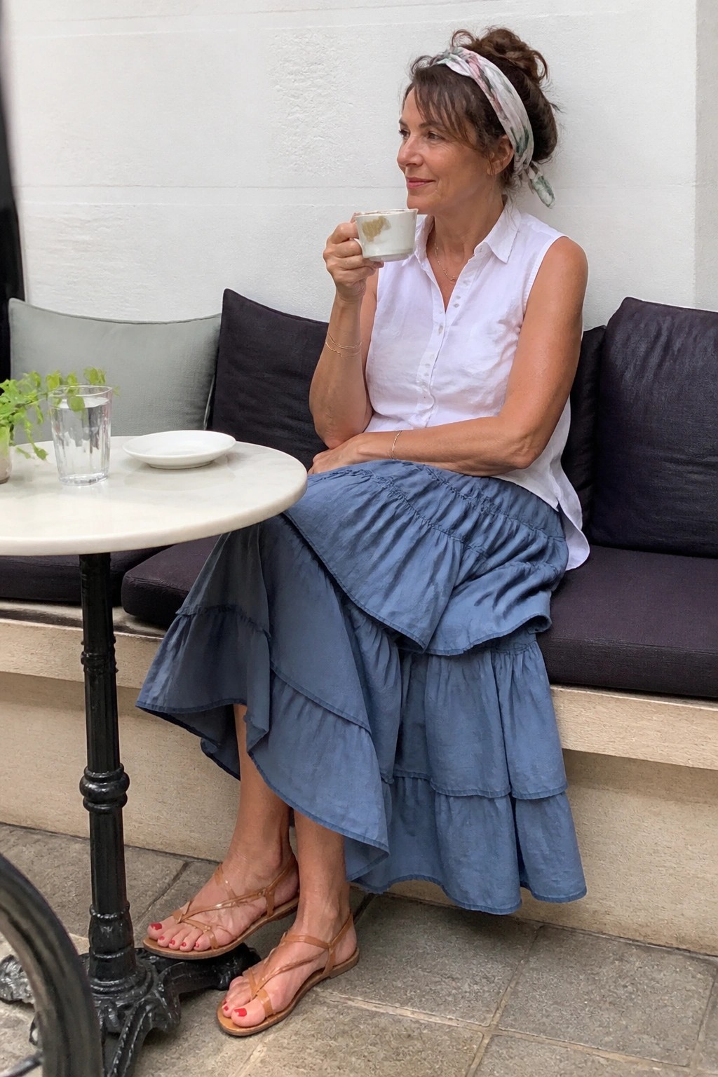 Woman in white sleeveless top and blue tiered skirt sips coffee outdoors