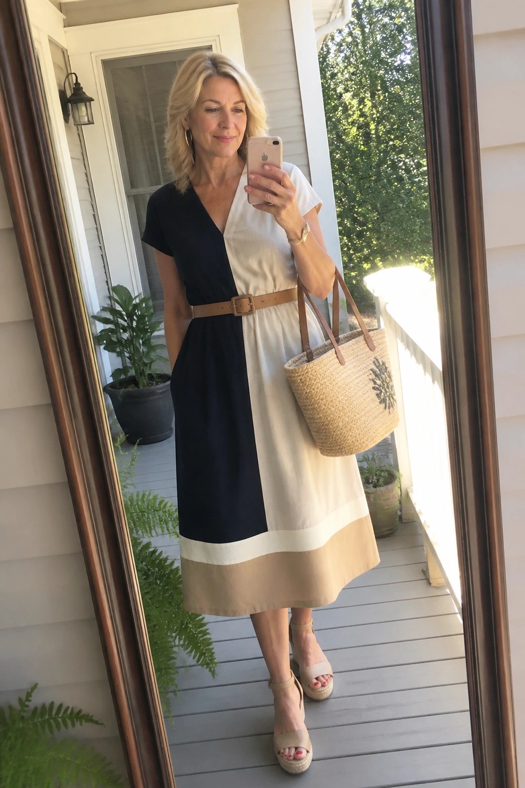 Woman over 50 in a black and cream colorblock midi dress with brown belt beige hem band wedge sandals and straw tote bag standing on porch.