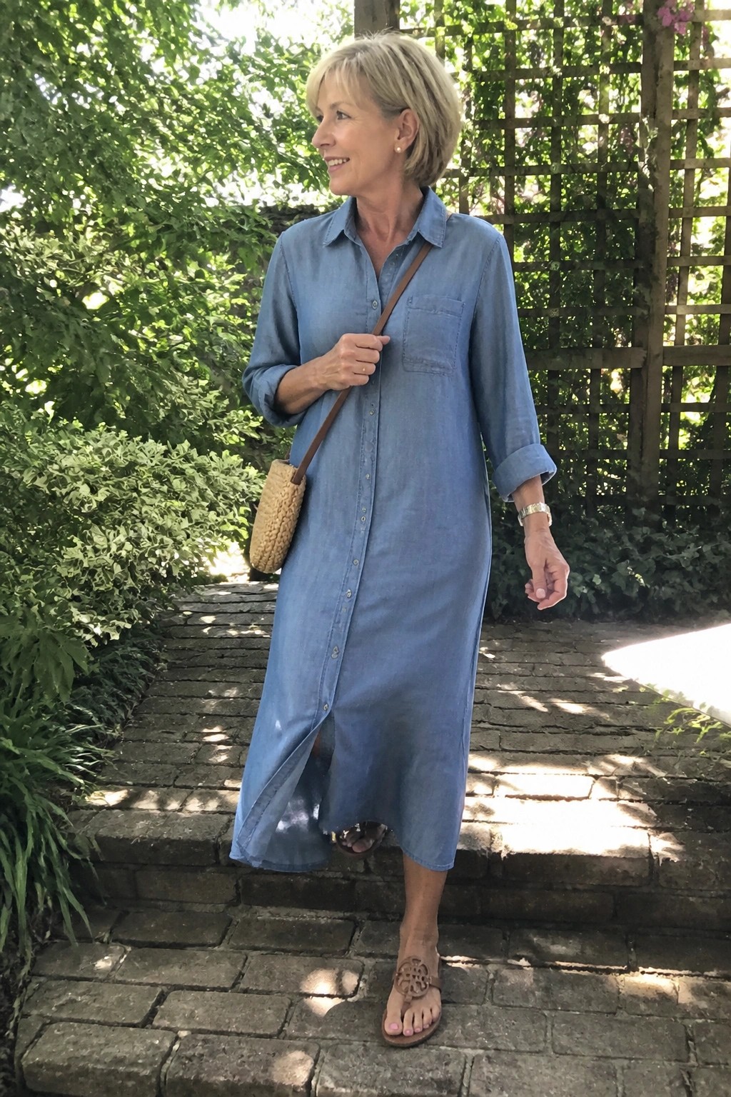 Woman in long blue denim shirt dress on brick steps with woven bag