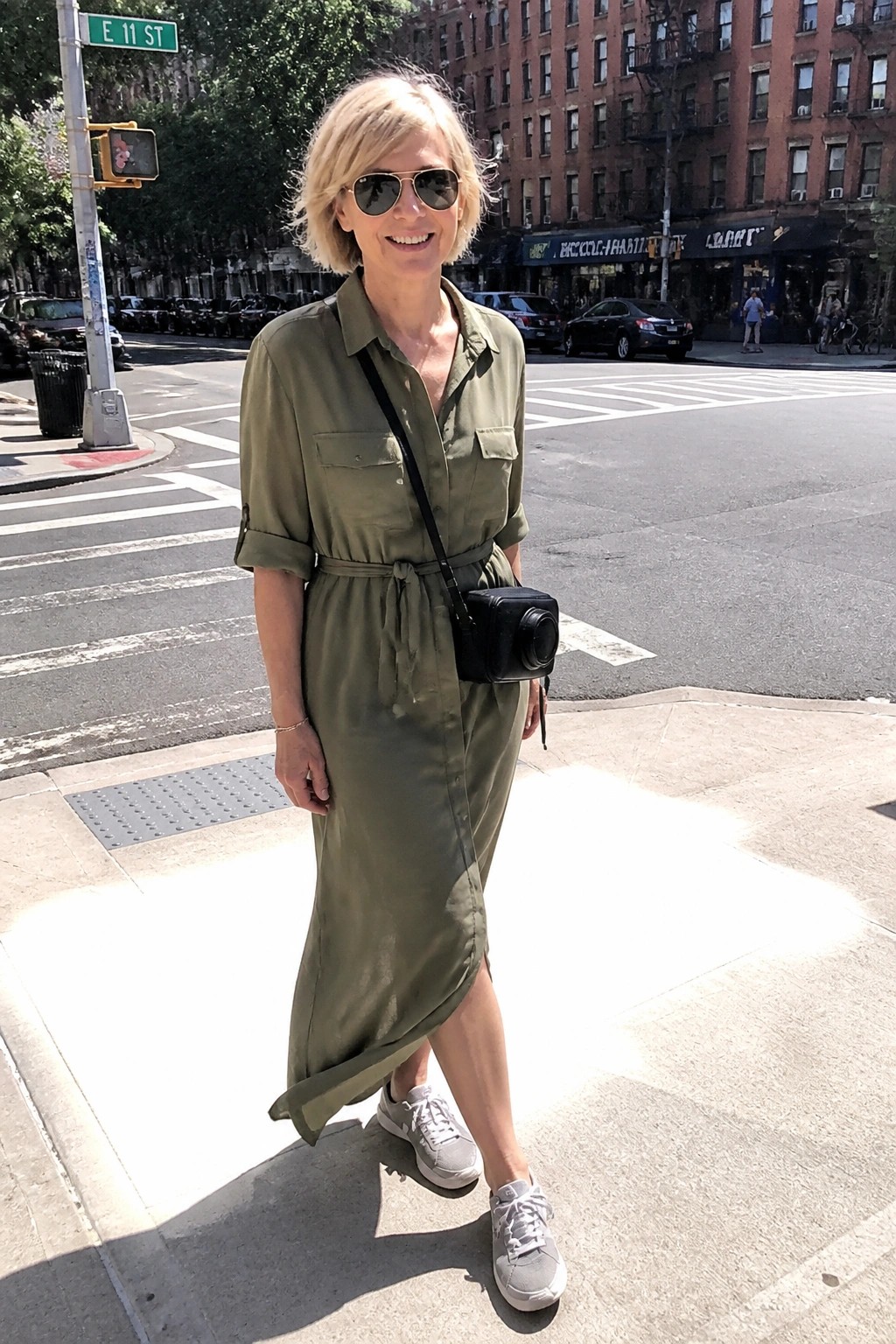 Smiling blonde woman in olive green shirt dress with camera on city street