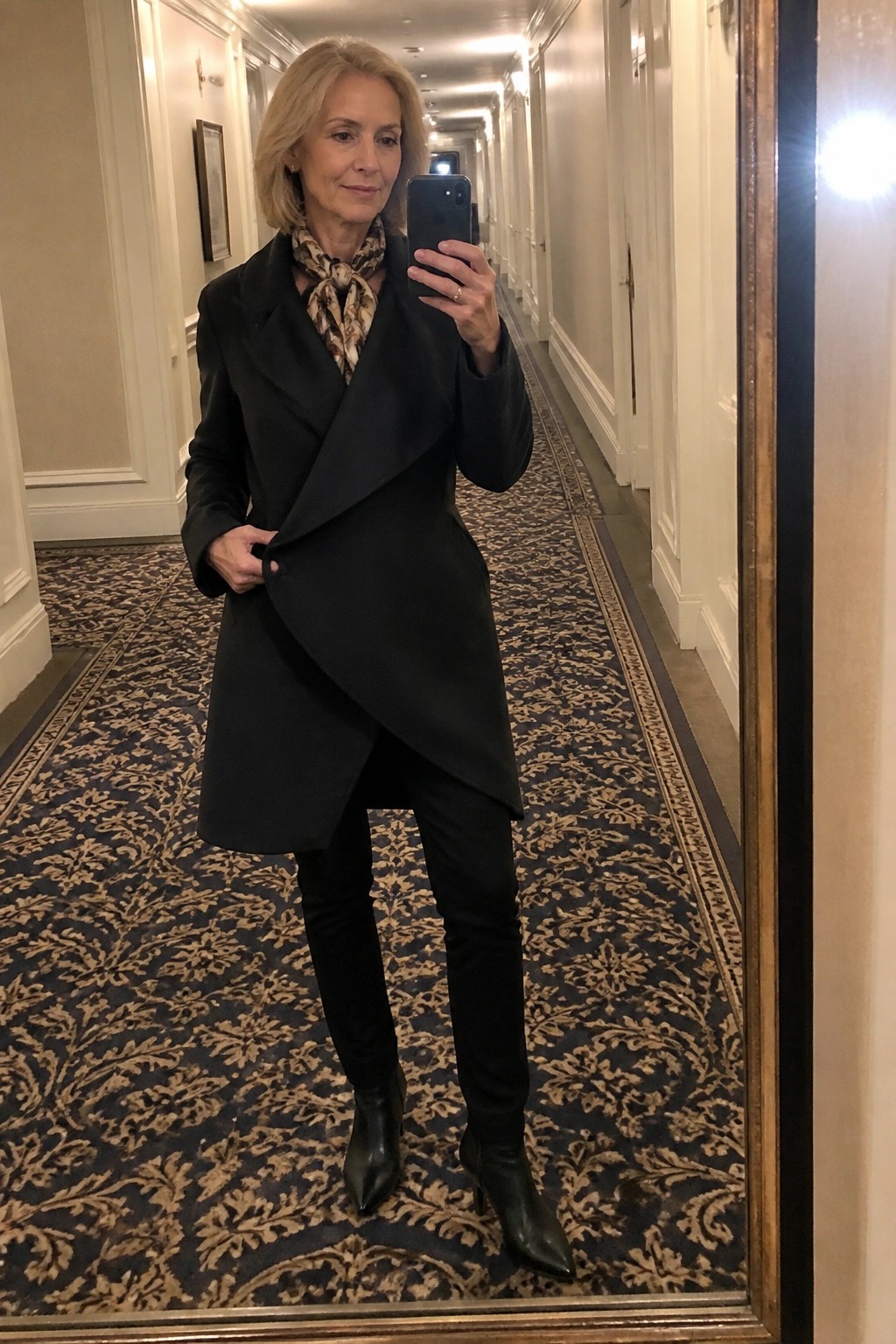 Blonde woman in black asymmetrical coat holding phone in elegant hallway mirror