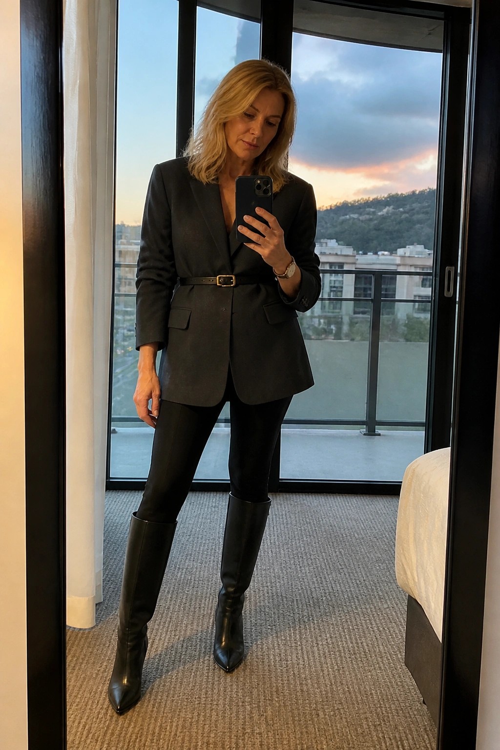 Blonde woman in black blazer and boots taking mirror selfie at dusk.