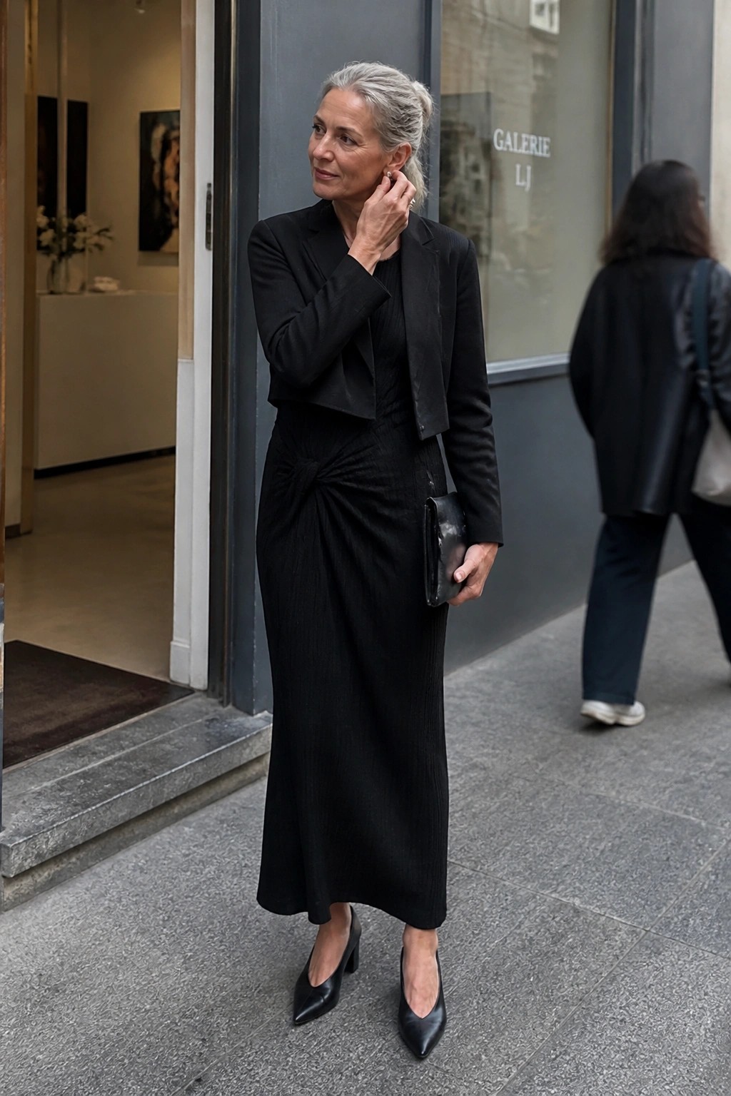 Woman with gray hair in black cropped blazer and knotted dress holding clutch on sidewalk