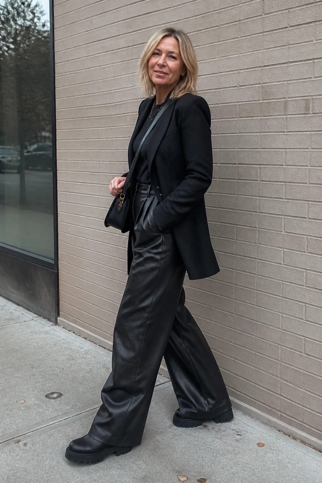 Blonde woman in black blazer and leather pants with bag against brick wall