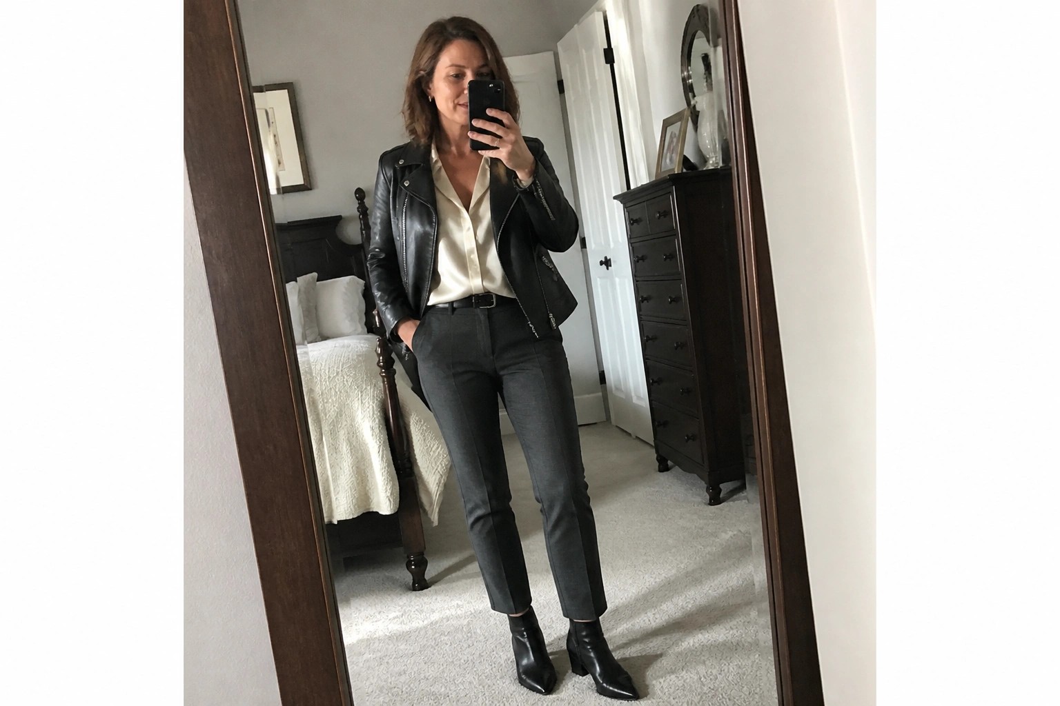 Woman wearing black leather moto jacket over white satin blouse with black trousers black belt and pointed black ankle boots in mirror selfie.