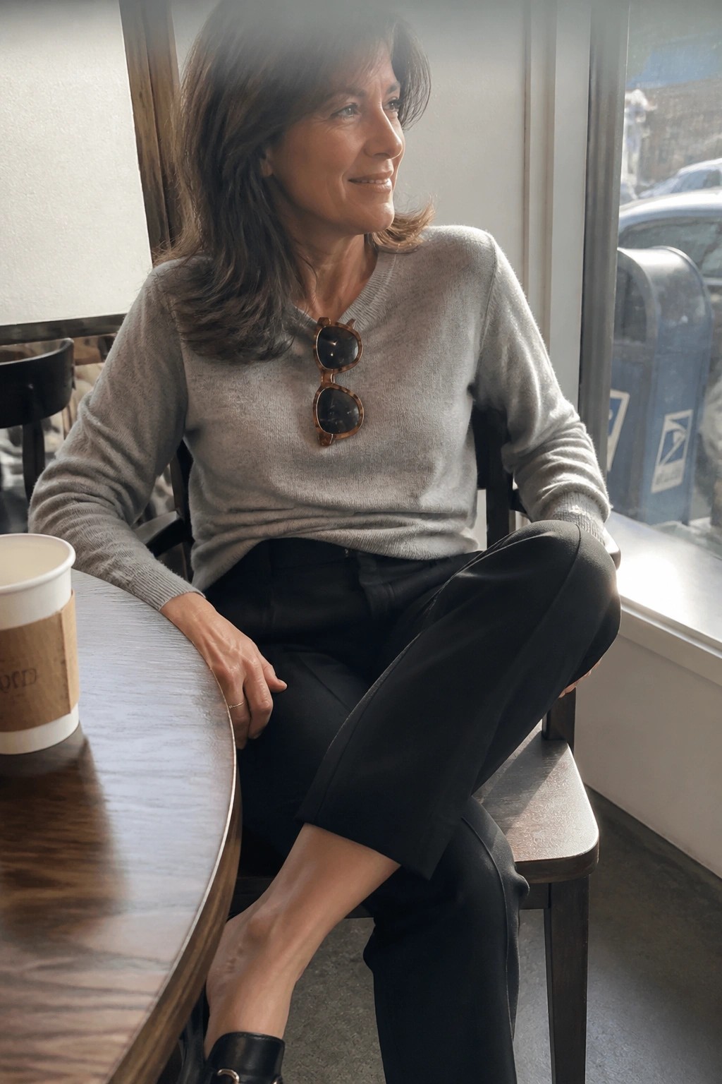 Smiling woman in gray sweater and black pants seated in cafe with coffee cup