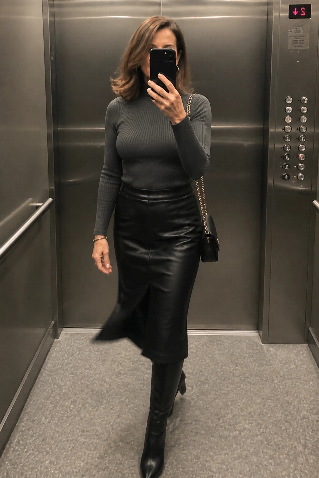 Woman in gray turtleneck, black leather skirt, and boots taking elevator selfie