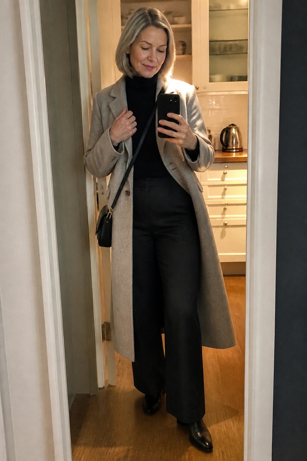 Woman with gray hair in long gray coat taking mirror selfie in kitchen doorway