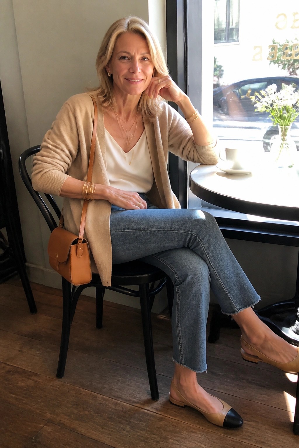 Blonde woman in beige cardigan and jeans smiling at sunny cafe window table