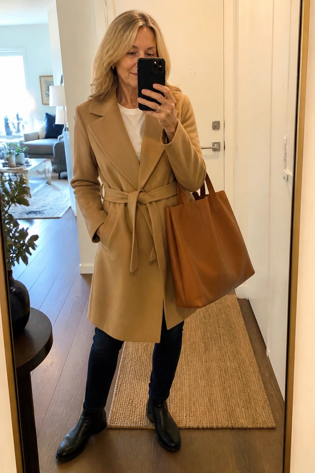 Woman over 50 in a camel belted wrap coat over a white top and dark jeans, paired with black ankle boots and a brown leather tote, shown in a mirror selfie