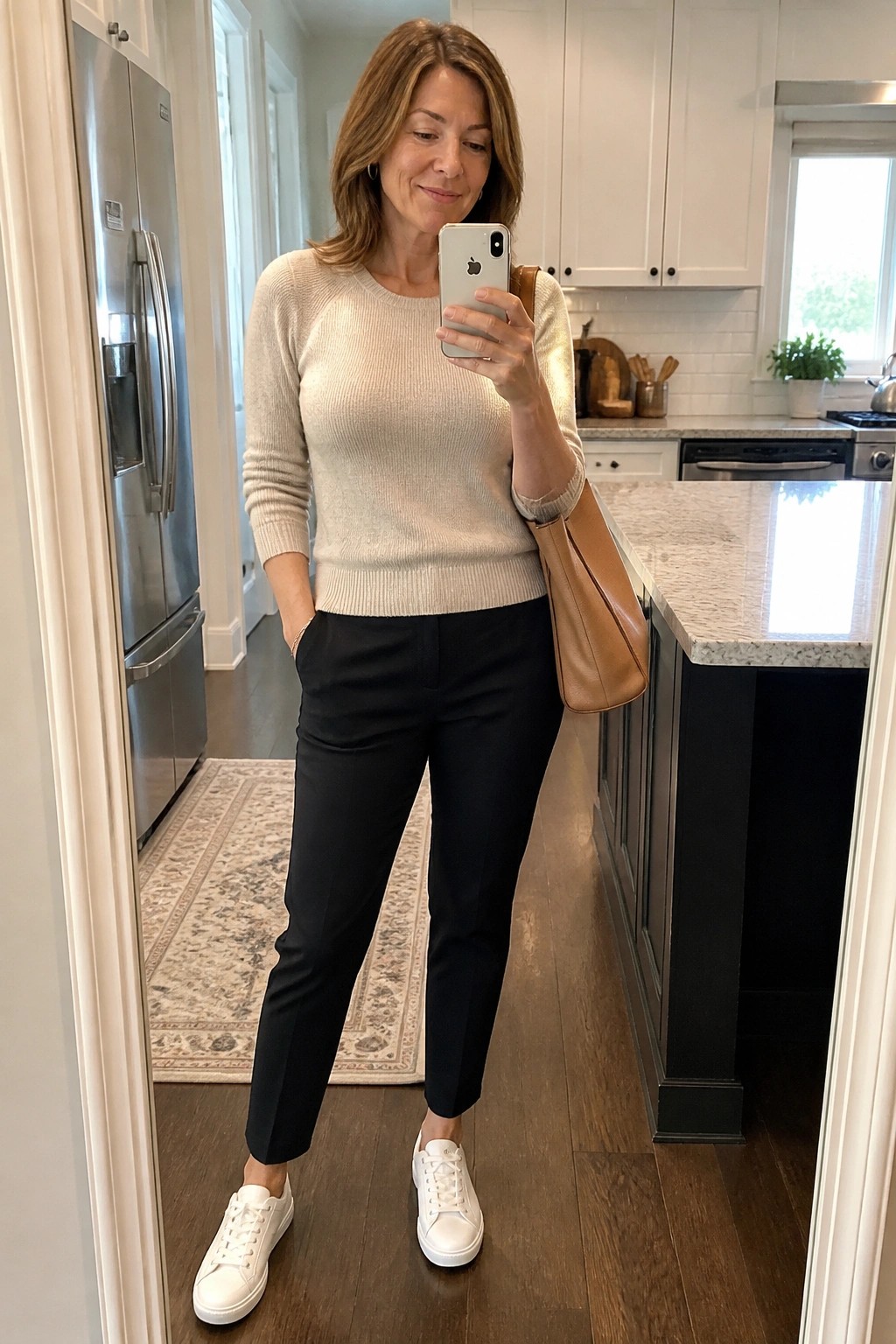Woman in beige sweater and black pants taking mirror selfie in kitchen