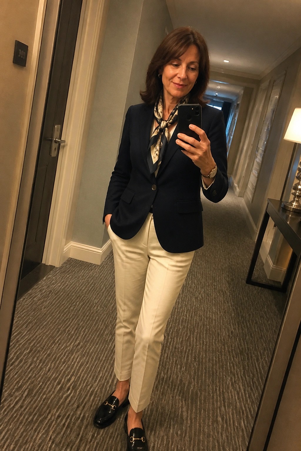 Woman in navy blazer and white pants taking mirror selfie in hallway
