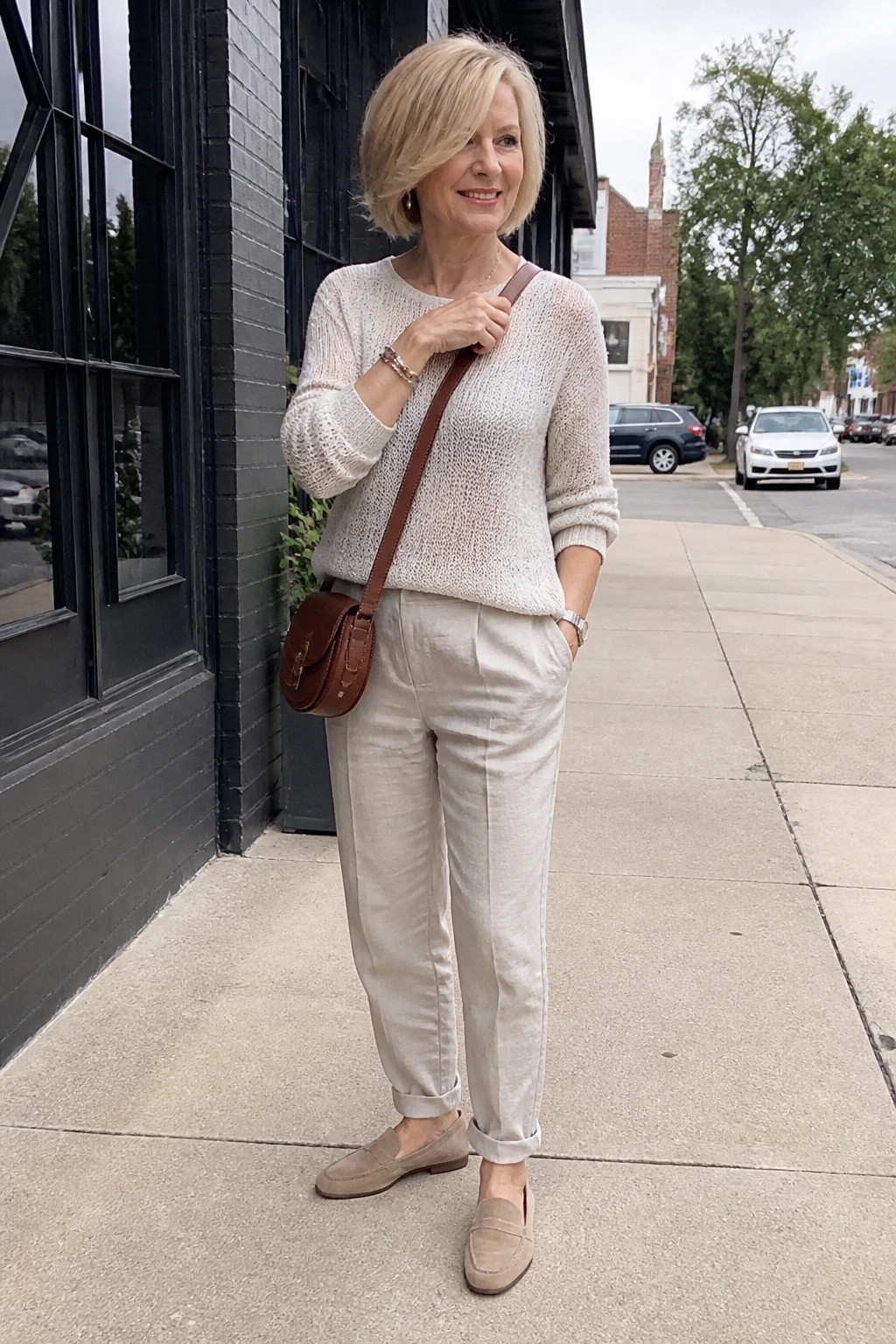 Blonde woman in cream knit sweater and beige pants with brown leather crossbody bag on sidewalk