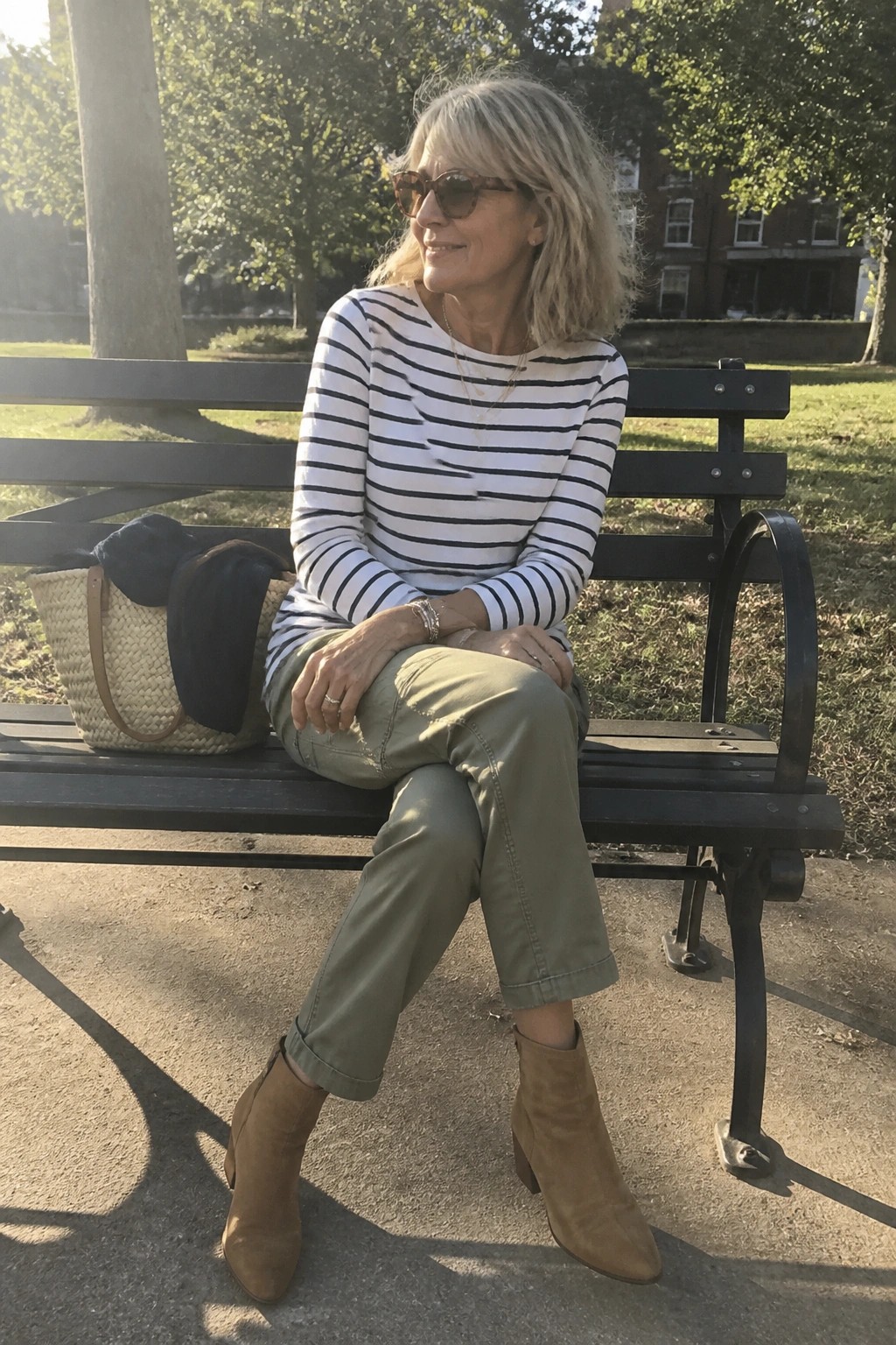 Woman in striped shirt and olive pants sits on park bench with straw bag