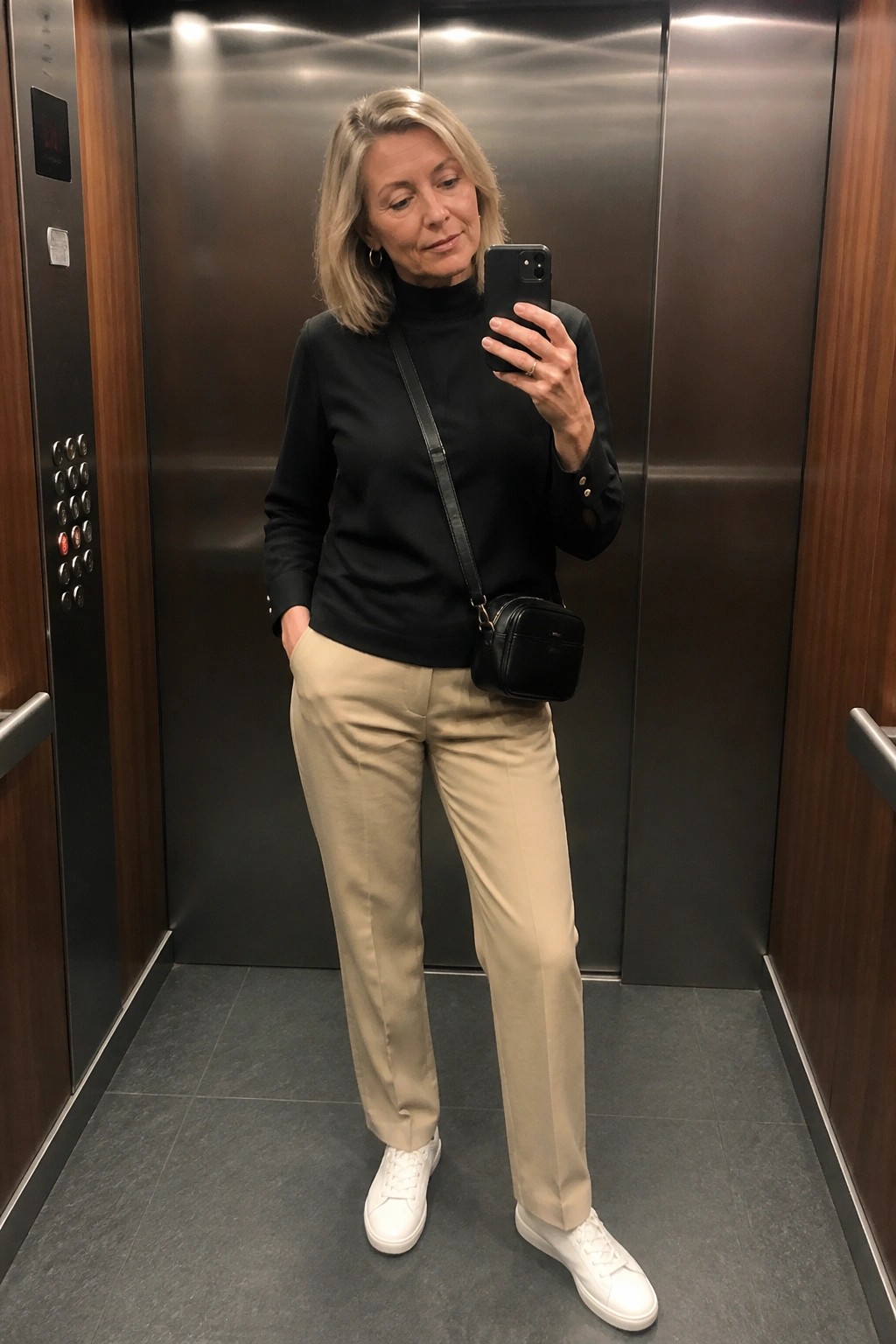 Blonde woman in black turtleneck and beige pants taking selfie in elevator with black bag