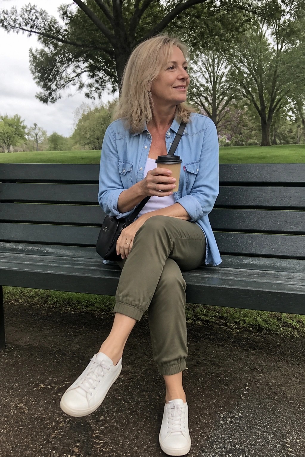 Blonde woman in blue denim shirt and olive pants holds coffee on park bench
